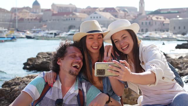 Three Young Friends Taking Selfies While Sitting On A Rock In Dubrovnik Bay