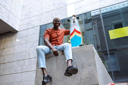 A low-angle shot of a  fashionable man with bleached hair holding his longboard while sitting on the wall