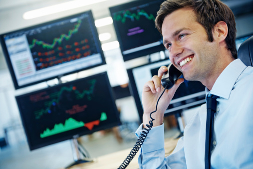 Profile shot of a smiling young businessman talking on the phone while sitting in front of monitors displaying financial information