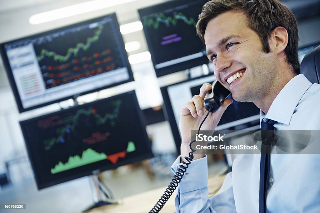 This has been a good financial quarter! Profile shot of a smiling young businessman talking on the phone while sitting in front of monitors displaying financial information Stock Market and Exchange Stock Photo