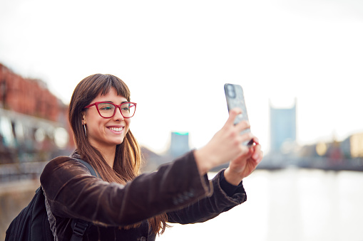 portrait beautiful caucasian young woman smiling taking selfie with her smart phone in the city street