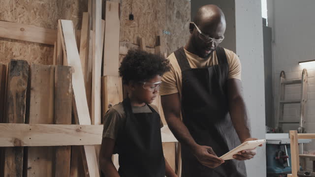 Dad Showing Blueprints on Tablet to Son in Carpentry Workshop