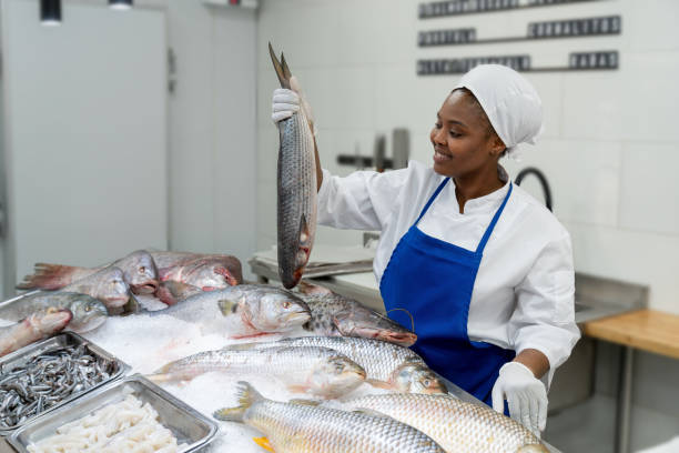 istock Black female fishmonger holding a fish from it's tail while smiling at the market stall 1607787868