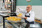 Caucasian mature manual worker at a seafood store serving paella in a plastic container for a delivery order