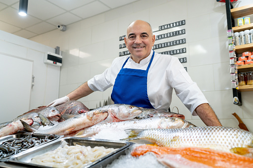 Mature male owner of a seafood market standing behind the seafood display counter while smiling at the camera - Fishmonger lifestyles