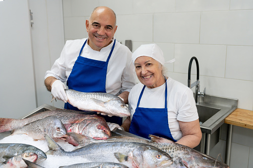 Happy senior fishmonger couple standing behind the fresh fish counter facing the camera smiling - Small business concepts