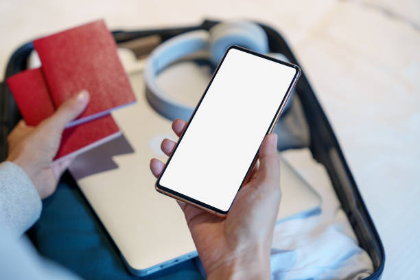 istock In a high-angle view, a woman is seen holding a mockup screen smartphone while packing a suitcase with various items including clothing, sunglasses, headphones, a laptop, and a passport. 1607796322