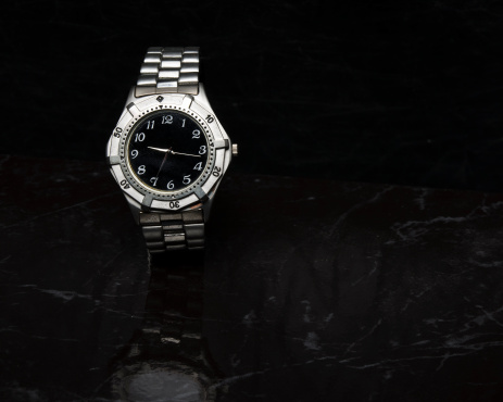A silver watch sits on black marble.  It has a reflection in the marble, and the background is black.  shot in studio.