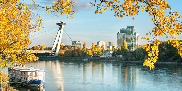 Danube river with old boat anchored on riverfront with modern office towers and Most SNP, popularly known as the UFO bridge, all framed by yellow autumn trees