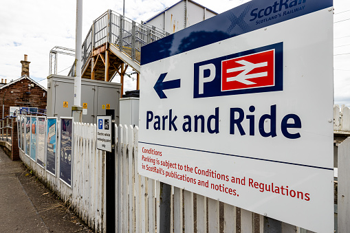 Close-up of a Park and ride sign at Cardross ScotRail train station, with a 'P' sign and arrow to parking, with a passenger bridge in the background