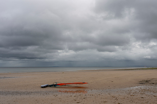 Langrune-Sur-Mer, France - 07 23 2023: A sailboard with his sail on the beach, the sea and a cloudy rainy sky behind