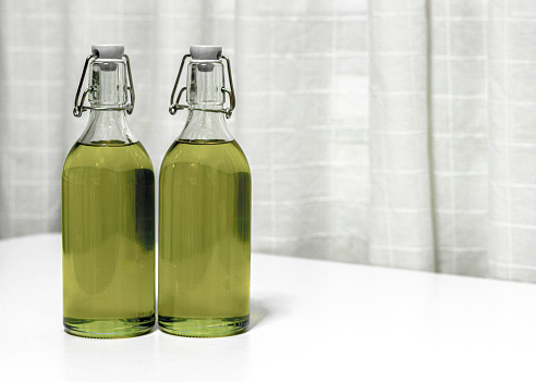 Extra virgin olive oil is bottled in glass with hermetic cap, with an olive green color, on the countertop of a kitchen table, there are fabric curtains behind it, with a white background.