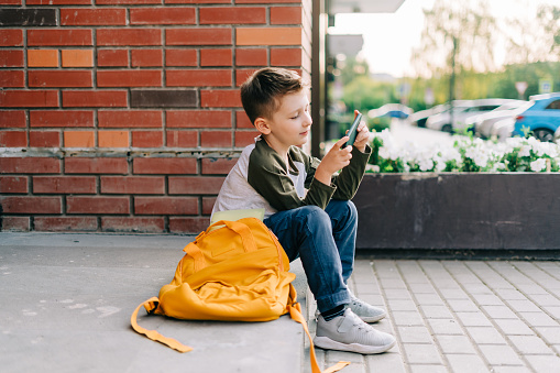 Back to school. Cute child with backpack, holding mobile phone, playing with cellphone. School boy pupil with bag. Elementary school student after classes. Kid sitting on stairs outdoors in the street