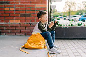 Back to school. Cute child with backpack, holding mobile phone, playing with cellphone. School boy pupil with bag. Elementary school student after classes. Kid sitting on stairs outdoor in the street