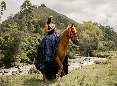 young woman with her quinceañera dress and tiara in her hair riding her horse outdoors along the river bank in the middle of nature.