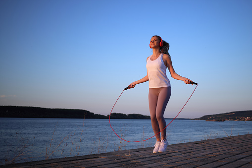 Beautiful young girl in her 20s with blonde hair by the river on a fitness path exercising with jumping rope while listening to music and enjoying the sunset. Photo taken in the golden hour