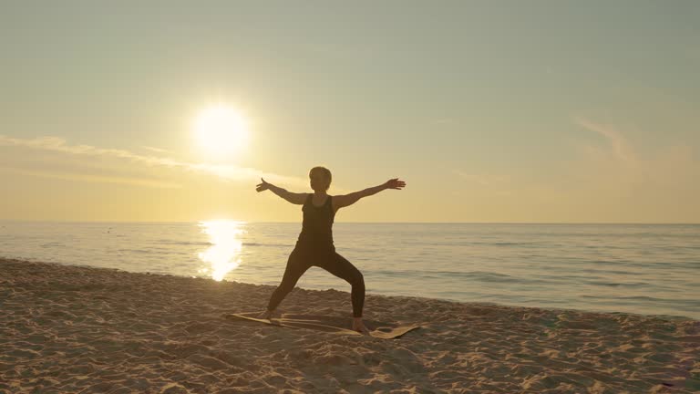 Sporty middle years woman standing in oriental position on mat doing yoga meditation and stretching at sunset beach