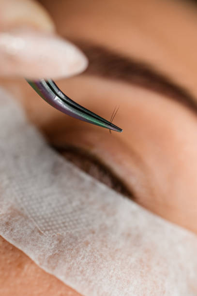 close-up of bunch of black false eyelashes in multi-colored steel tweezers. Eyelash extension procedure. close-up of bunch of black false eyelashes in multi-colored steel tweezers. Blurred face of female client in the background. Eyelash extension procedure. clump eyelashes stock pictures, royalty-free photos & images