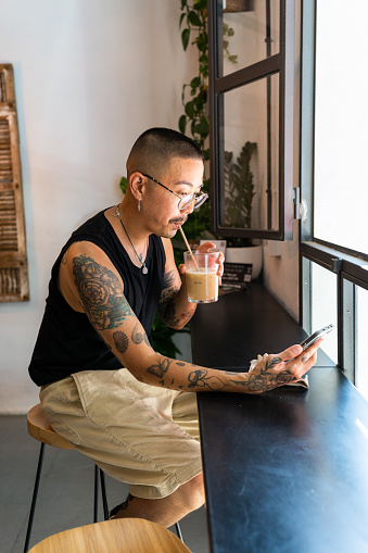 Vertical photo of a gay man drinking iced coffee and using phone sitting in a cafeteria