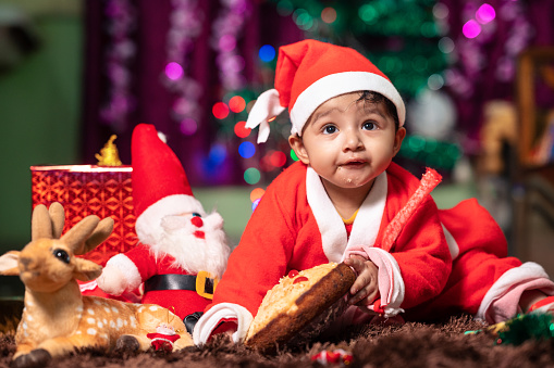 Cute little baby boy in Santa hat and clothes lying down on the carpet on the floor in front of the Christmas tree and a Santa doll,  Concept of Christmas and Holiday with little toddler,