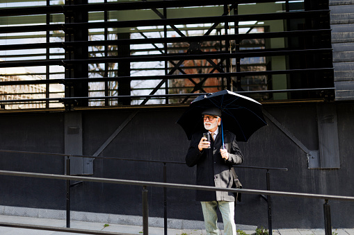 A senior businessman walking and holding an umbrella and drinking coffee