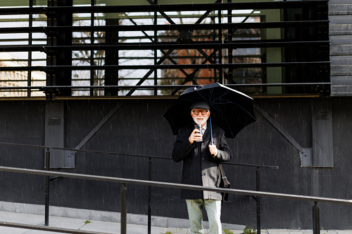 A senior businessman walking and holding an umbrella and drinking coffee