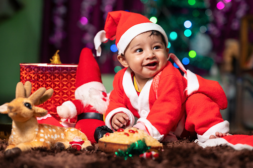 Happy Smiling little toddler boy dressed in Santa dress and red Christmas hat on Christmas decorated background with Santa doll and some gift boxes, Celebrating Christmas .