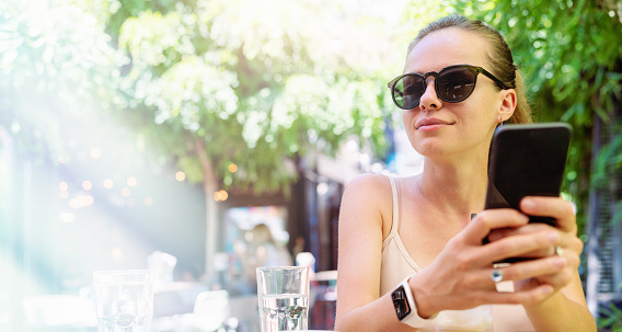 Young urban woman wearing sunglasses sits at the table in outdoor summer cafe holding smart phone in her hands and smiling.
