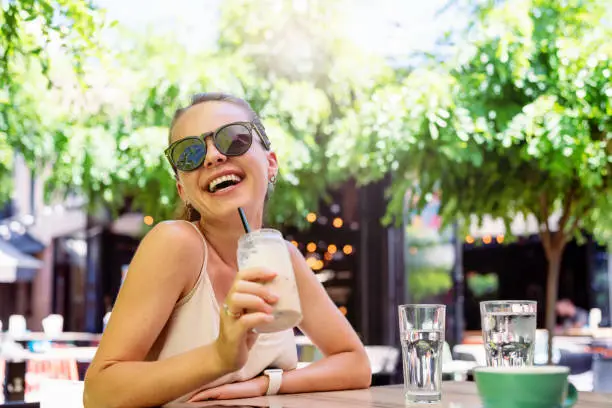 Happy young brunette woman sitting in the open-air cafe drinking iced-coffee and laughing.