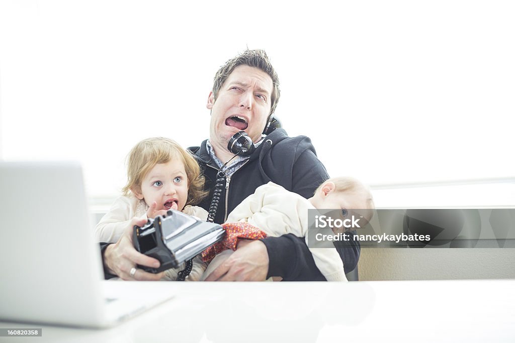 Humorous Dad in Office Trying to Multitask Work and Parenting A dad tries to multitask in the office with work and parenting and finds it frustrating.  The dad is sitting in his office holding two young daughters and talking on the phone with his laptop open on his desk. Humor Stock Photo