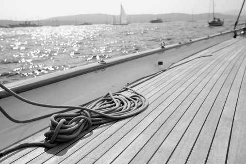 Black and White image of a classic sailing yacht teak deck and coiled rope. Other yachts in distance. Depth of field with focal point on foreground.
