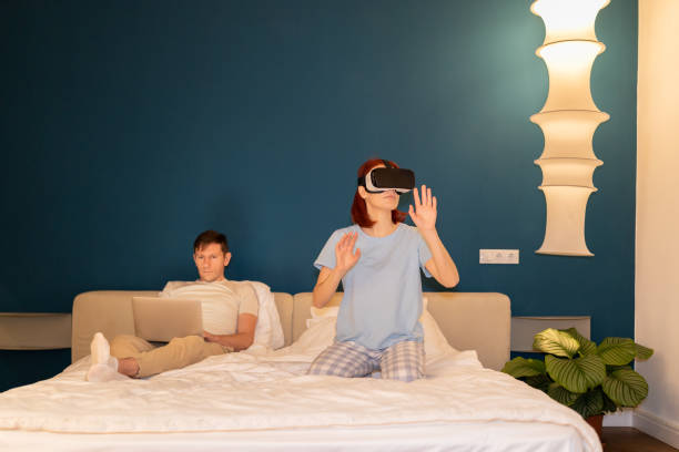 istock Woman in vr glasses immersive in game while man with laptop work in bed different spend time at home 1608247712