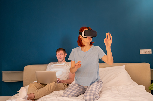 Interested woman in glasses virtual reality sits on bed with husband manages vr goggles from laptop. Young female in 3D headset exploring cyberspace metaverse world touching unreal things playing game