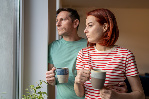 Upset girl and man hold cup of coffee, look out window, sad and anxious. Family problems. Unhappy husband and wife hustle about life and problem solving. Feelings loneliness in couple