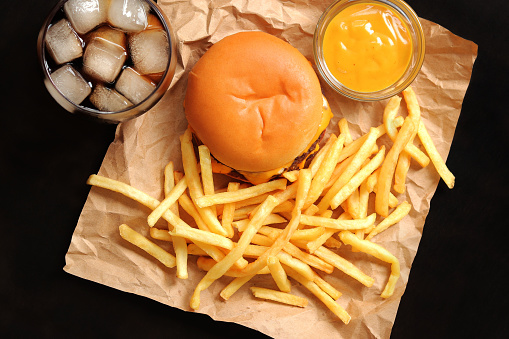 Cheeseburger and french fries on brown paper and wooden table, top view. Burger, french fries, cola with ice in a glass and mustard. Fast food