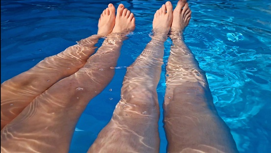 Seen over the legs of a mother and her daughter sitting side by side in turquoise pool water