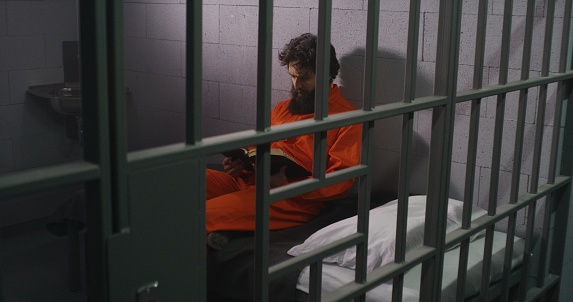 Male prisoner in orange uniform sits on bed, reads Bible, prays, looks at barred window in prison cell. Inmate serves imprisonment term for crime in jail. Correctional facility. Faith in God concept.