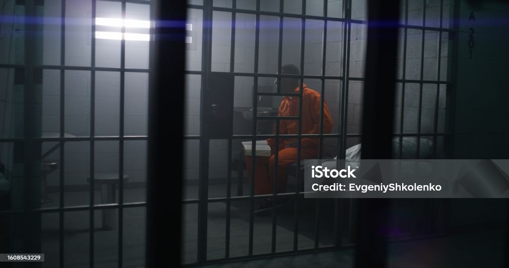 African American prisoner sits on the bed, reads Bible African American prisoner in orange uniform sits on the bed, reads Bible in prison cell. Male criminal serves imprisonment term for crime in jail or detention center. Faith in God. View through bars. Criminal Stock Photo