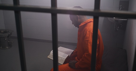 African American prisoner in orange uniform sits on bed behind bars, reads Bible in prison cell. Male criminal serves imprisonment term for crime in jail. Detention center or correctional facility.