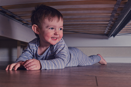 Toddler baby crawls and hides under the bed. Child climbed under the sofa and sits on the floor. Kid boy age one year eight months, full length