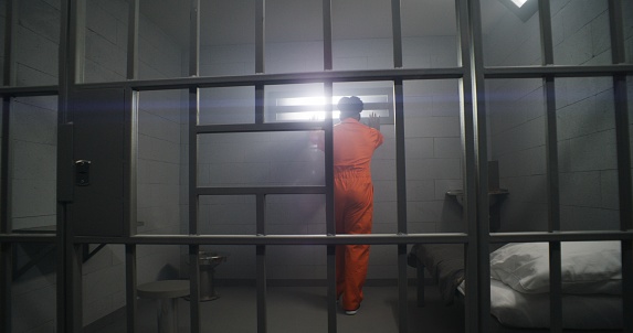 African American prisoner in orange uniform looks at barred window and leans on prison cell bars. Depressed inmates in detention center. Murderers serve imprisonment term in jail cell.