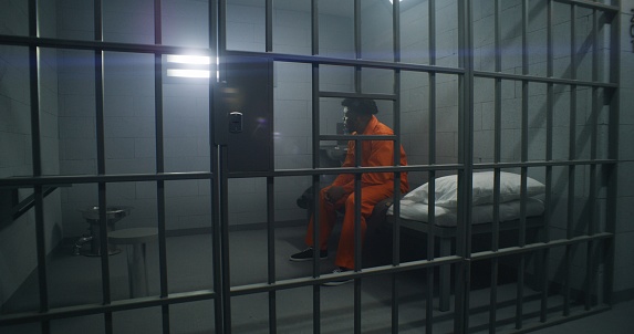 African American inmates in detention center or correctional facility. Depressed man in orange uniform sits on prison bed and looks at barred window. Prisoners serve imprisonment term in jail cell.