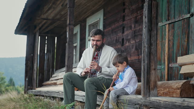Little boy with dad playing on woodwind wooden flutes - ukrainian sopilka. Family duet, folk music concept. Father and son in traditional embroidered shirts. They sitting on porch of old wooden house