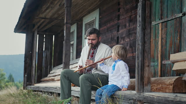Little boy with dad playing on woodwind wooden flutes - ukrainian sopilka. Family duet, folk music concept. Father and son in traditional embroidered shirts. They sitting on porch of old wooden house