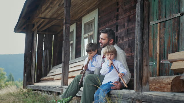 Little boys with dad playing on woodwind wooden flutes - ukrainian sopilka. Family, folk music concept. Father and sons in traditional embroidered shirts. They sitting on porch of old wooden house