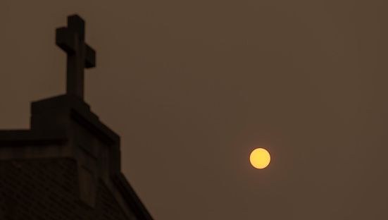 Church cathedral roof with cross in the dark sky with sun is under the smog from Canadian fires in Jim Thorpe (Mauch Chunk) of Appalachian ountains, Poconos, Pennsylvania. Low angle view