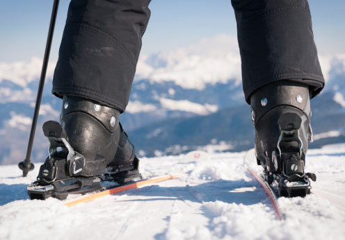 Close-up on the boots and bindings of a skier about to set off down a piste, with snowcapped mountains in the background.