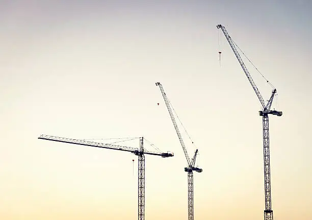 Three large construction cranes at a large industrial building site, photographed at dawn.