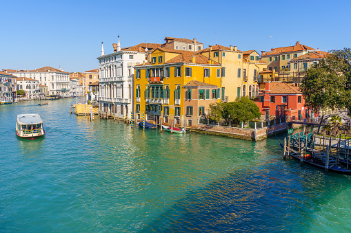 Pedestrian walkway leading to stone paved pier, tour boat going on water surface of canal between residential buildings in Venice, Italy