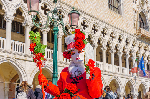 Venice, VE, Italy - February 13, 2024: masquerade person with red hat and ancient palace on the background during carnival